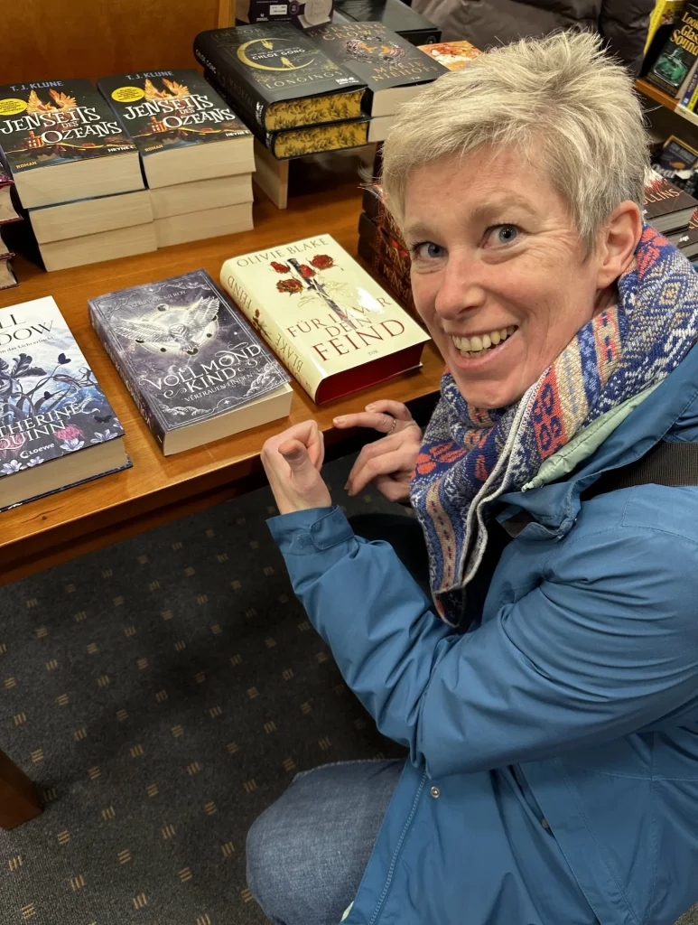 Die Autorin Kerry McKilroy lächelt in einer Buchhandlung neben einem Büchertisch, auf dem ihr Fantasy-Roman „Vollmondkind – Vertrauen finden“ ausliegt.