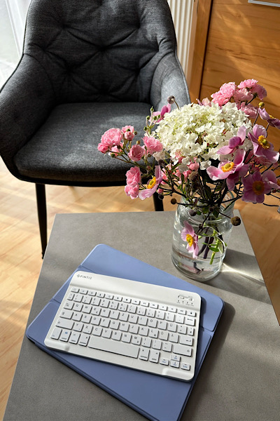 Ein grauer Sessel, davor ein Tisch mit einem Tablet, einer Tastatur und einer Blumenvase, mit rosa und weißen Blumen darin, die Sonne scheint schräg auf das Bild und liefert Schattierungen