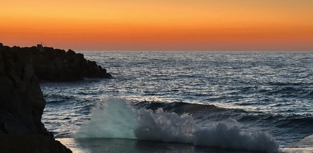 Sonnenuntergang am Meer, links sind Felsen, am vorderen Bildrand schlagen Wellen an einen Steg