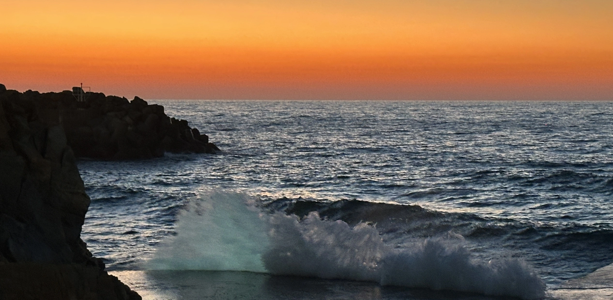 Sonnenuntergang am Meer, links sind Felsen, am vorderen Bildrand schlagen Wellen an einen Steg