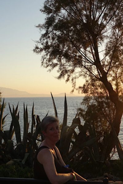 untergehende Sonne am Meer, eine Bank im Vordergrund, auf der eine Frau sitzt und sich nach hinten zum Fotografen wendet,